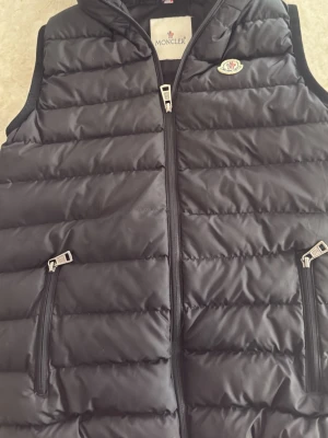 Svart dunväst från Moncler - Snygg svart dunväst från Moncler med quiltad design och två dragkedjeförsedda fickor framtill. Västen har en klassisk Moncler-logga på bröstet och innerfoder med serietidningsmönster. Perfekt för lager-på-lager och streetstyle.