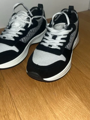 Dior grå och svarta sneakers - Snygga sneakers från Dior i svart och grått med vit sula. Ovandelen har mesh och mockadetaljer samt Dior-logga på sidan. Skorna har vita skosnören och en chunky sula som ger en modern look.