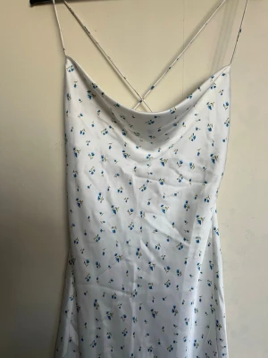 Vit blommig klänning med smala band - Säljer en vit klänning med små blå blommor och tunna axelband som korsas i ryggen. Klänningen har en lös passform, öppen rygg och ett mjukt, svepande fall. Perfekt för sommarens varma dagar!🥰🥰