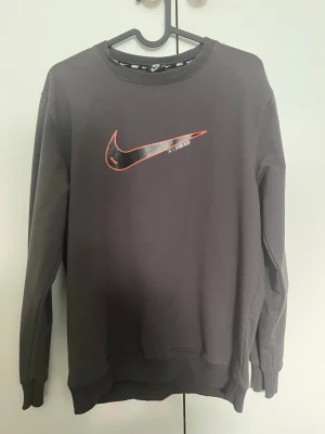 Mörkgrå Nike sweatshirt med logga - Säljer en mörkgrå sweatshirt från Nike i storlek M. Tröjan har en stor Nike-logga i rött och svart på bröstet och ribbade muddar vid ärmslut och nederkant. Perfekt för en sportig och avslappnad stil.