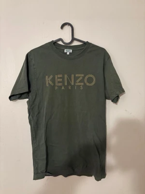 Grön Kenzo Paris t-shirt - Snygg grön t-shirt från Kenzo Paris med stort guldfärgat logotryck på bröstet. Klassisk passform med rund hals och korta ärmar. Perfekt för dig som gillar streetwear och vill ha något stilrent men ändå med en twist.