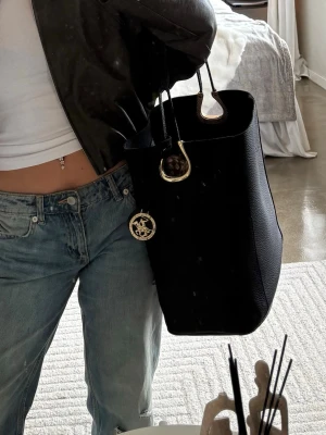 Svart Beverly Hills Polo Club bucket bag - Snygg svart bucket bag från  Beverly Hills Polo Club i strukturerat läder. Väskan har guldfärgade metalldetaljer. Praktiska handtag och rymlig form som rymmer allt du behöver. Perfekt accessoar för en trendig look. Avtagningsbart innerfack och ett knäppe så du kan stänga väskan. Har en liten skada längst ner i hörnet men inget man märker av.