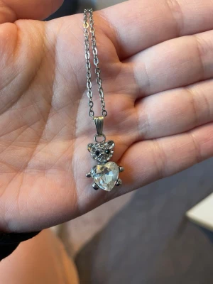 Halsband med björn och hjärtformad sten - Sött halsband i silverfärg med en björnberlock dekorerad med små klara stenar och en stor hjärtformad kristall som kropp. Kedjan är tunn och stilren, perfekt för att ge en gullig touch till din outfit.