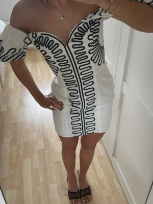 Vit kortklänning med svarta detaljer - Supersnygg vit kortklänning med offshoulder och puffärmar. Klänningen har svarta broderade mönster framtill och längs ärmarna, samt en djup v-ringning🌸