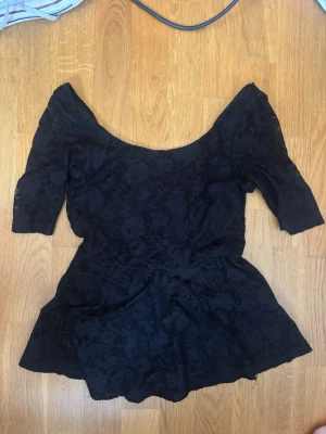 Svart spetsblus med offshoulder - Snygg svart blus i spets med offshoulder och trekvartsärm. Blusen har en figurnära passform upptill och faller ut nedtill för en feminin siluett. Perfekt att styla med jeans eller kjol för en trendig look.
