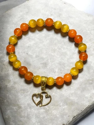Armband med orange och gula pärlor - Snyggt armband med runda pärlor i orange och gult, dekorerat med guldfärgade detaljer och ett hängsmycke i form av två hjärtan. Perfekt accessoar för att addera färg till din stil. Elastiskt band för enkel påtagning.