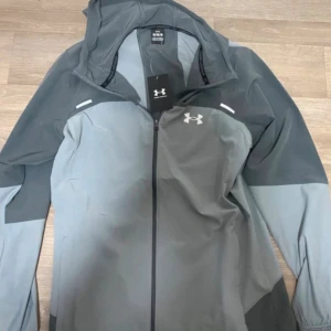 Under Armour - Snygg grå träningsjacka med huva från Under Armour. Jackan har dragkedja framtill, logga på bröstet och reflekterande detaljer. Tillverkad i ett lätt och funktionellt material som passar perfekt till löpning eller gym. Stora Under Armour-trycket på ryggen.