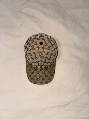 Beige keps från Gucci med GG-mönster - Säljer en beige keps från Gucci med det klassiska GG-monogrammet över hela kepsen. Kepsen har böjd skärm och en liten knapp på toppen. Materialet är canvas med detaljer i brunt och svart. Perfekt för dig som vill ha en stilren och lyxig accessoar. Frakt ingår.