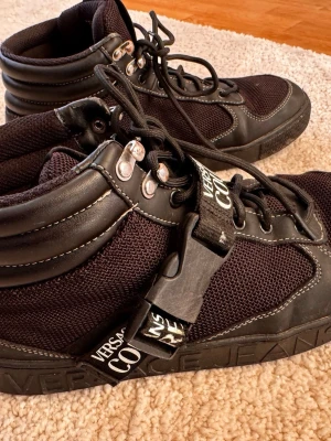 Svarta Versace Jeans Couture sneakers - Snygga svarta sneakers från Versace Jeans Couture med högt skaft, detaljer i både mesh och läderimitation samt vita kontrastsömmar. Skorna har snörning, metallöglor och en bred rem med logga över vristen. Tydlig logotyp på sidan och plös.