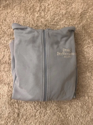 Ljusblå hoodie Peak Performance - Ljusblå hoodie från Peak Performance med dragkedja framtill och broderad logga på bröstet. Hoodien har korta ärmar, två fickor fram och ribbade muddar. Perfekt för en avslappnad och sportig stil. Materialet känns mjukt och bekvämt.