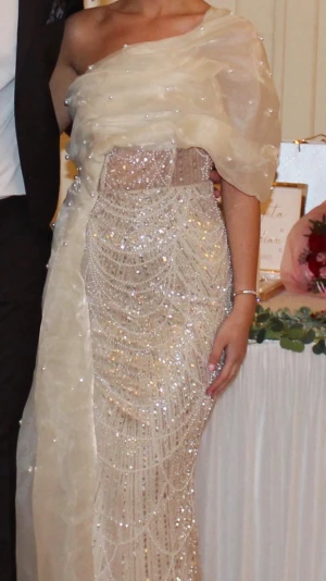 Glittrig långklänning med pärlor - Säljer en elegant långklänning i champagnefärg med glittriga paljetter och pärldetaljer. Klänningen har ett axelbandslöst snitt med ett draperat tyg över ena axeln och en figurnära passform. Perfekt för bal eller festliga tillfällen.