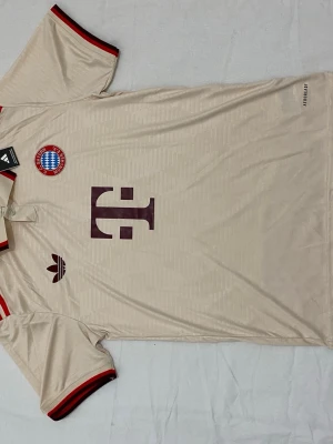 En Bayern München bortatröja 24/25 - Prisen kan diskuteras. Snygg beige Bayern München matchtröja från Adidas med klubbmärke och Adidas-logga på bröstet. Tröjan har röda och svarta detaljer på krage och ärmslut samt stort sponsortryck framtill. Tillverkad i lätt och ventilerande material, perfekt för fotboll eller träning.