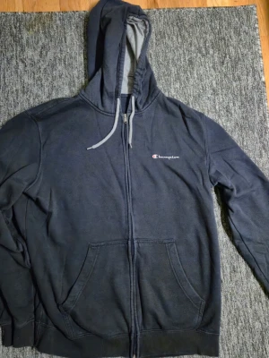 Svart zip hoodie från Champion XL - Säljer en svart zip hoodie från Champion med dragsko i huvan och två stora fickor framtill. Champion-loggan är broderad på bröstet. Hoodien är tillverkad i mjukt bomullsmaterial och har ribbade muddar vid ärmslut och nederkant. Storlek XL, skickas eller så hämtas i hässelby