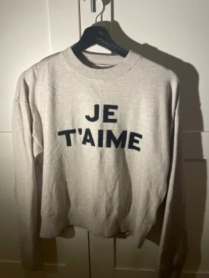 Beige sweatshirt från Gina Tricot - Beige sweatshirt från Gina Tricot med svart text 'JE T'AIME' framtill. Tröjan har rund halsringning, långa ärmar och ribbade muddar vid hals, ärmslut och nederkant. Perfekt för en avslappnad och trendig look.