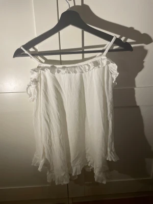 Vit offshoulder blus med volang - Säljer en vit offshoulder blus med tunna axelband och långa, vida ärmar. Blusen har volangdetaljer vid halsringning, ärmslut och nederkant. Materialet är lätt och krinklat, vilket ger en avslappnad och luftig känsla.