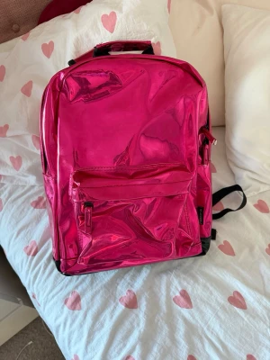 Metallic rosa ryggsäck - Säljer en riktigt snygg metallic rosa ryggsäck med en stor ytterficka och justerbara axelband. Väskan har en glansig finish och svarta detaljer på baksidan och axelremmarna. Perfekt för dig som vill sticka ut och gillar färg!