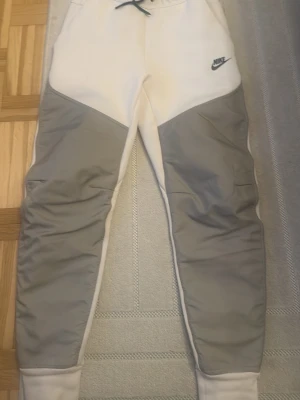 Nike mjukisbyxor i vitt och grått - Säljer ett par snygga mjukisbyxor från Nike i vitt med gråa paneler på benen. Byxorna har dragsko i midjan, muddar vid bensluten och en liten Nike-logga på vänster lår. Perfekta för chill dagar eller träning. Materialet känns mjukt och bekvämt.