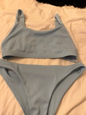 Ljusblå ribbad bikini - Säljer en ljusblå bikini från Shein med ribbad struktur. Överdelen har breda axelband och en enkel, sportig form med justerbara band och spänne i ryggen. Underdelen är klassiskt skuren och matchar toppen perfekt. Perfekt för strandhäng!