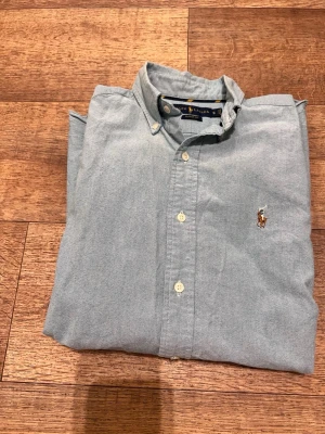 Ljusblå skjorta från Ralph Lauren - Klassisk ljusblå skjorta från Ralph Lauren med button-down krage och broderad logga på bröstet. Skjortan har normal passform och vita knappar framtill. Tillverkad i mjuk bomull som känns skön mot huden.