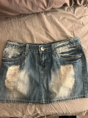 Blå jeanskjol med slitningar - Säljer en blå jeanskjol i kort modell med slitna detaljer och råa kanter framtill. Kjolen har klassiska fem fickor, bälteshällor och knappstängning. Perfekt för dig som gillar en avslappnad och trendig stil.