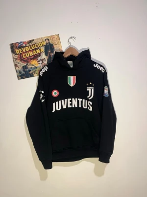 Juventus svart hoodie med tryck - Snygg svart hoodie med Juventus-logga, klubbmärke och sponsortryck från Jeep på axlarna. Stora vita bokstäver över bröstet och flera detaljerade emblem. Perfekt för dig som älskar fotboll och vill representera ditt lag med stil.