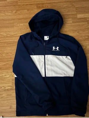 Marinblå vindjacka från Under Armour - Snygg marinblå vindjacka från Under Armour med vit diagonal panel över bröstet och dragkedja framtill. Jackan har huva, två sidofickor och logga på bröstet. Perfekt för blåsiga dagar och sportiga outfits.