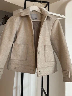 Beige ullrock från 157 - Stilren beige ullrock från 157 med klassisk krage, stora framfickor och knappar vid ärmslut och nederkant. Rocken har en rak passform och är helfodrad för extra komfort. Perfekt för dig som gillar minimalistisk och tidlös stil.