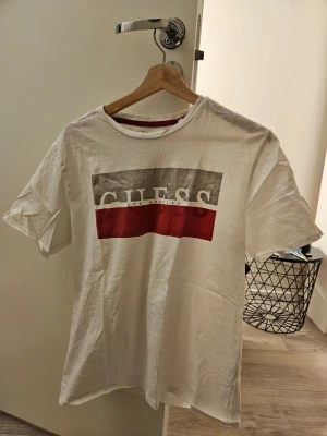 Vit GUESS t-shirt  - Säljer en vit t-shirt från GUESS med stort tryck i grått och rött på bröstet där det står 'GUESS Los Angeles'. Storleken på etiketten är XL, men enligt min mening passar den bättre för storlek L.