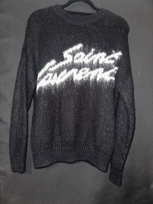 Svart stickad Saint Laurent-tröja - Stickad svart tröja från Saint Laurent med vit logotyp i grafisk stil över bröstet. Tröjan har rund hals, långa ärmar och ribbstickade muddar vid ärmslut och nederkant. Materialet ser ut att vara mjukt och lite fluffigt.