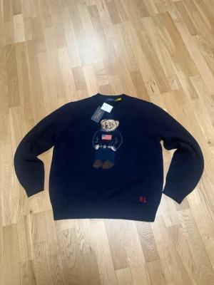 Mörkblå stickad tröja Polo  - Mörkblå stickad tröja från Polo Ralph Lauren med den ikoniska Polo Bear-björnen på bröstet, klädd i amerikansk flaggtröja. Rund halsringning och ribbade muddar. RL-broderi nertill. Perfekt för dig som gillar klassisk preppy stil. L men passar mer M