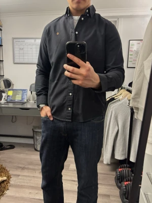 Svart skjorta  - Stilren svart skjorta  med klassisk button-down krage och vita knappar. Skjortan har en liten guldfärgad ankare-brodyr på bröstet och är tillverkad i mjuk bomull. Perfekt till jeans eller chinos för en clean look. OBS! har et litet hål mer går inte igenom, se sista bild