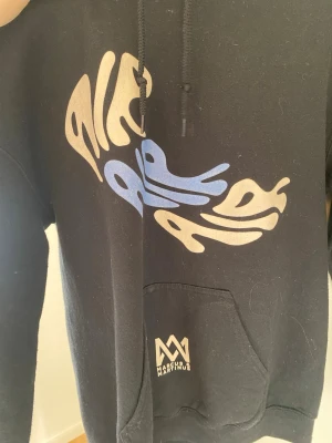 Svart hoodie från Marcus & Martinus - Cool svart hoodie med stor grafisk print i beige och blått på bröstet. Klassisk huva med snören och stor magficka framtill. Materialet är mjukt och bekvämt, perfekt för att chilla eller hänga med kompisar. Logotypen finns på fickan.