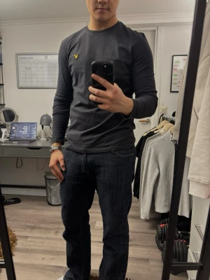 Svart långärmad tröja Lyle & Scott - Snygg svart långärmad tröja från Lyle & Scott med klassisk gul logga på bröstet. Tröjan har rund halsringning och är gjord i mjukt bomullsmaterial. Perfekt för en clean och stilren look.