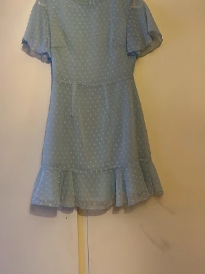 Ljusblå prickig kortklänning - Söt ljusblå kortklänning med vita prickar och volangkant nedtill. Klänningen har korta, fladdriga ärmar och rund halsringning. Perfekt för sommarens alla tillfällen och ger en romantisk vibe.