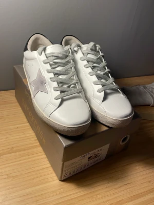 Golden Goose vita sneakers med stjärna - Snygga vita sneakers från Golden Goose med grå stjärna på sidan, grå skosnören och svart detalj på hälen. Skorna är i läder och har en klassisk låg profil med vintage-inspirerad sula. Perfekta för dig som gillar streetstyle och exklusiva märken. Kan gå ner i pris vid snabb affär!!
