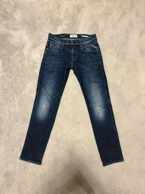 Mörkblå slim fit jeans från Replay - Snygga mörkblå slim fit jeans från Replay med klassisk femficksmodell och subtila slitningar på låren. Jeansen har normal midja, dragkedjegylf och kontrastsömmar. Perfekta för dig som gillar en modern och stilren look.