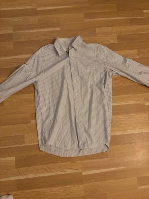 Vit och blå randig skjorta från Arket - Snygg vit skjorta med tunna blå ränder och relaxed fit. Skjortan har klassisk krage, knäppning framtill och långa ärmar. Perfekt för en chill och stilren look.