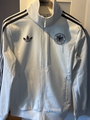 Vit Adidas track jacket Tyskland - Vit track jacket från Adidas med svarta ränder på ärmarna och Tysklands fotbollsförbunds logga på bröstet. Dragkedja framtill, hög krage och två fickor med dragkedja. Klassisk retrostil med Adidas Trefoil-logga. Perfekt för sport eller streetwear.