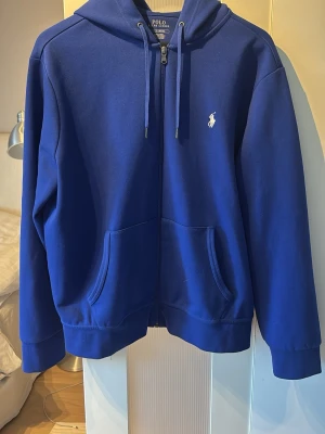 Blå hoodie från Polo Ralph Lauren - Snygg blå hoodie från Polo Ralph Lauren med dragkedja, två fickor framtill och klassisk huva med dragsko. 