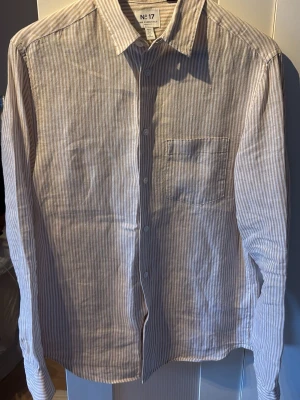 Randig skjorta från H&M Essentials - Säljer en beige och vit randig skjorta från H&M Essentials, modell No 17. Skjortan har klassisk krage, bröstficka och knappar framtill. Tillverkad i bomull och har lång ärm. Perfekt för en clean och avslappnad stil.