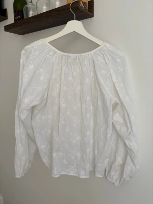 Vit blommig blus med knytning - Söt vit blus med diskret blommönster och rynkad halsringning. Blusen har långa ärmar och knytning i halsen för en avslappnad och romantisk look. Perfekt att styla med jeans eller kjol för en trendig vibe. Ej tvättat än därav hur den ser ut på framsidan - men den tvättas innan jag skickar! 