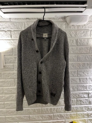 Grå stickad kofta från H&M - Mysig grå stickad kofta från H&M med sjalkrage och knäppning framtill. Koftan har långa ärmar och ribbade muddar. Perfekt för lager-på-lager och chill dagar.