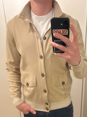 Beige cardigan från Gran Sasso - Snygg beige cardigan med ribbstickad krage och muddar. Koftan har stora fickor framtill med knappar och stängs med knappar hela vägen. Perfekt för lager-på-lager och passar till jeans eller chinos. Helt ny. Nypris 3999kr