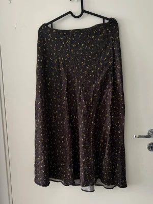 Svart långkjol  - Svart långkjol med små gula blommor. Kjolen har en lätt utsvängd form och faller snyggt längs benen. Perfekt för dig som gillar en feminin och stilren look. Materialet känns mjukt och svalt, troligen polyester eller viskos.