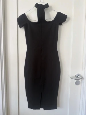 Svart offshoulder klänning med slits - Stilren svart klänning med offshoulder-design och hög hals. Klänningen har en figurnära passform, slits baktill och dragkedja i ryggen. Perfekt för dig som gillar en modern och elegant look.