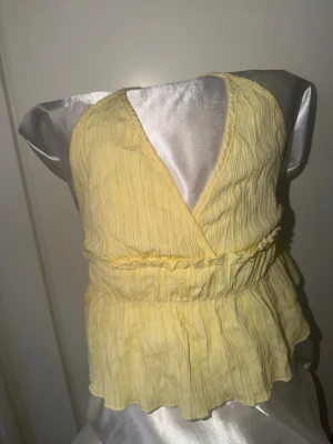 Gul omlott linnetopp med knyt - Söt ljusgul omlottopp i linnematerial med knyt i ryggen och volang nertill. Toppen är ärmlös och har en v-ringad halsringning, perfekt för varma dagar och en trendig look.
