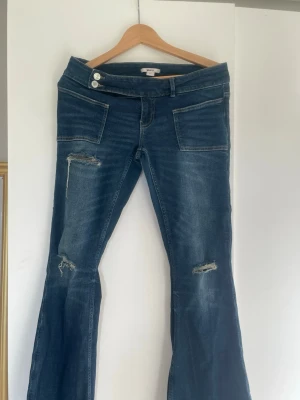 Blå bootcut jeans med slitningar - Snygga blå bootcut jeans med slitningar på benen och två knappar i midjan. Jeansen har framfickor och bakfickor, samt en lätt tvättad look. Perfekta för dig som gillar en avslappnad och trendig stil.