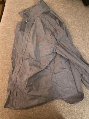 Blå skjorta från Ralph Lauren - Klassisk blå skjorta från Ralph Lauren med orange broderad logga på bröstet. Skjortan har knappar framtill och lång ärm. Perfekt för en stilren och avslappnad look.
