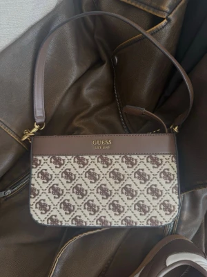 Brun Guess-väska - Säljer denna superfina bruna Guessväskan. Den är i princip helt oanvänd, men har något gult vid sömnen inuti väskan som jag tror är lim. Annars superfin! 
