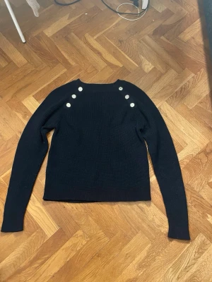 Svart stickad tröja med knappar - Snygg svart stickad tröja med långa ärmar och dekorativa silverfärgade knappar på axlarna. Tröjan har en rak passform och rund halsringning. Aldrig använd. 